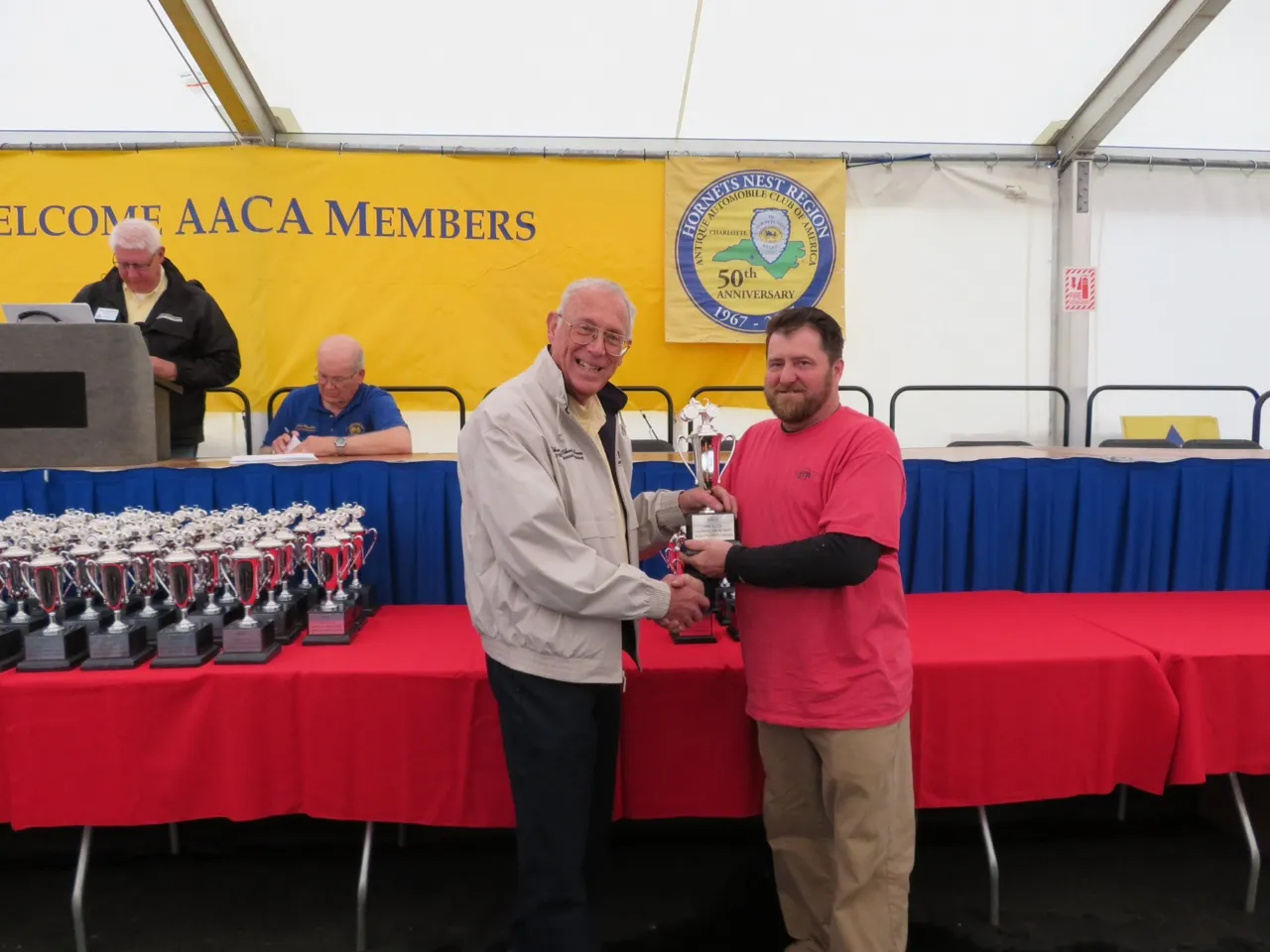 A man in a red shirt receiving a trophy beneath a “Welcome AACA Members” banner, a table of trophies alongside.