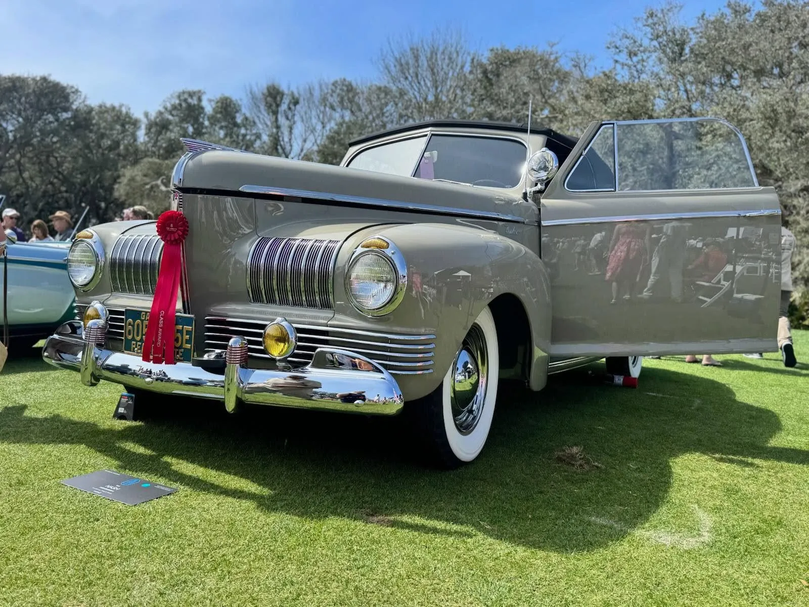 A khaki classic convertible on a show-field lawn with both doors open and a red ribbon on its grille.