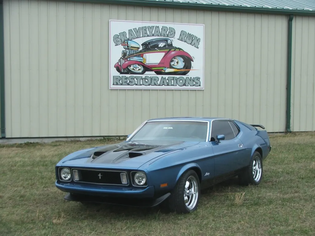 Restored 1973 Ford Mustang Mach 1 in blue, front three-quarter, under the hand-painted Graveyard Run shop sign.
