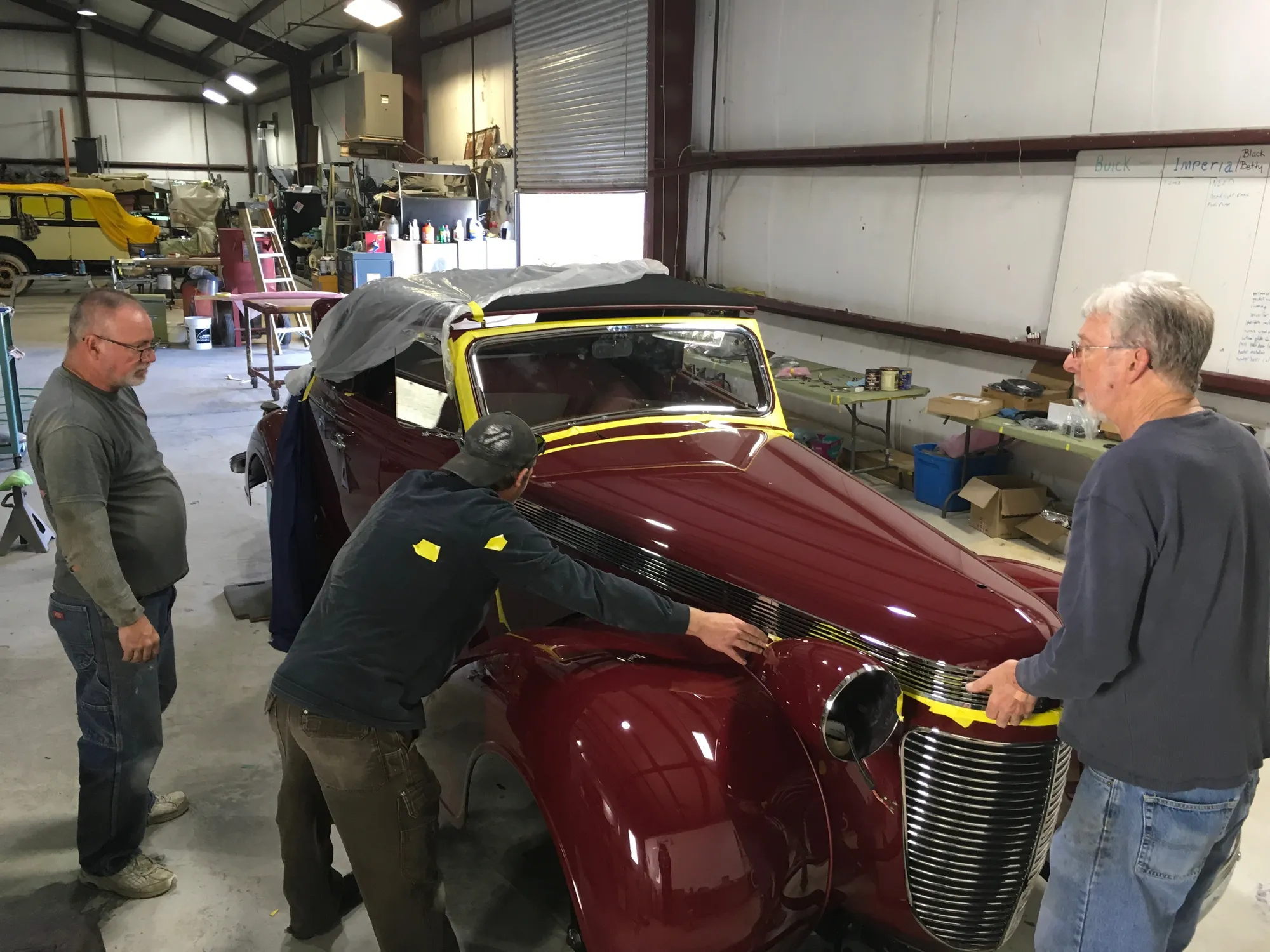 A restorer's hands fitting polished chrome trim to a bare-metal classic body under shop tungsten light
