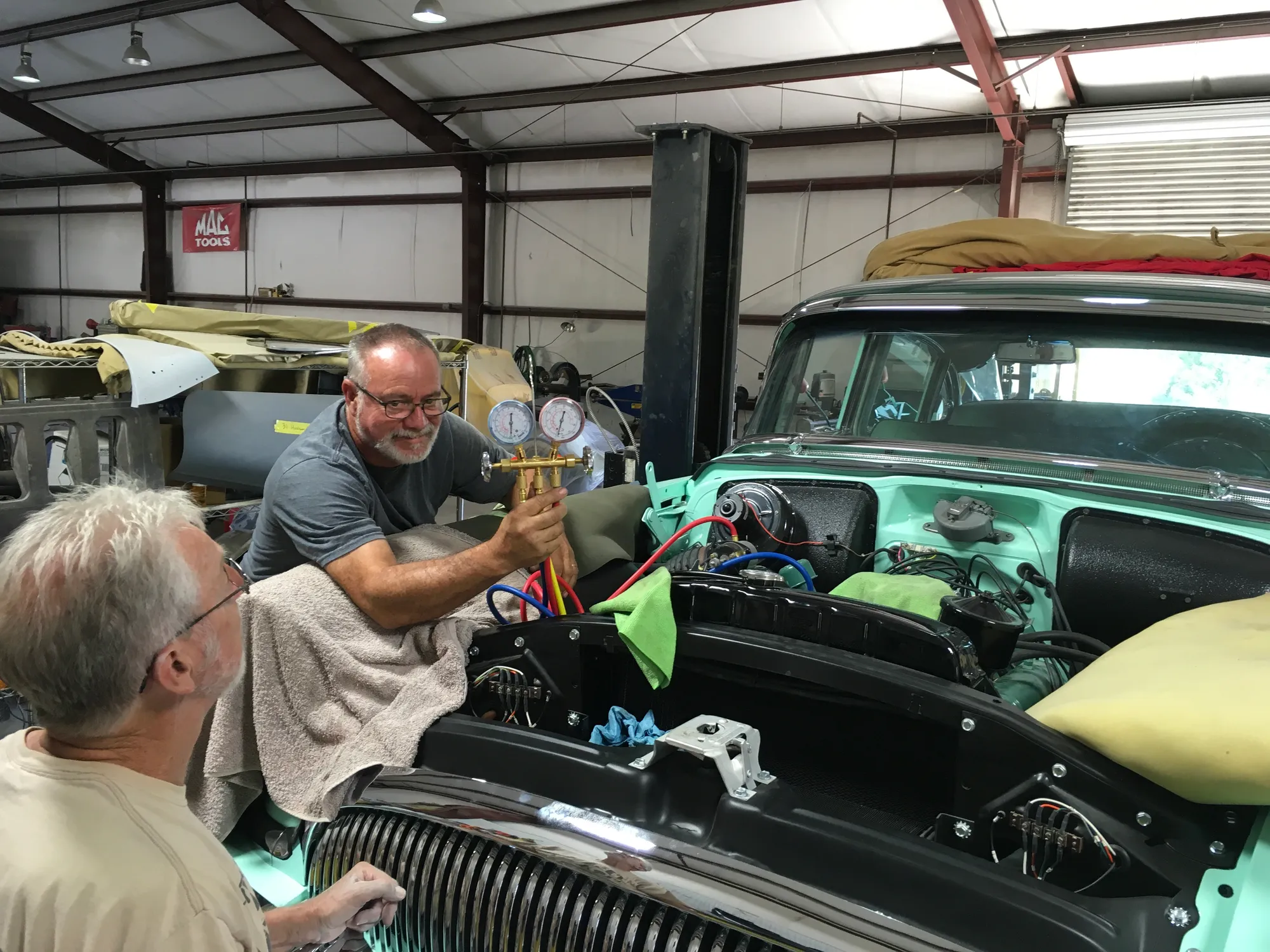 Two men at the open hood of a seafoam-green classic in the shop, one holding a set of A/C manifold gauges.