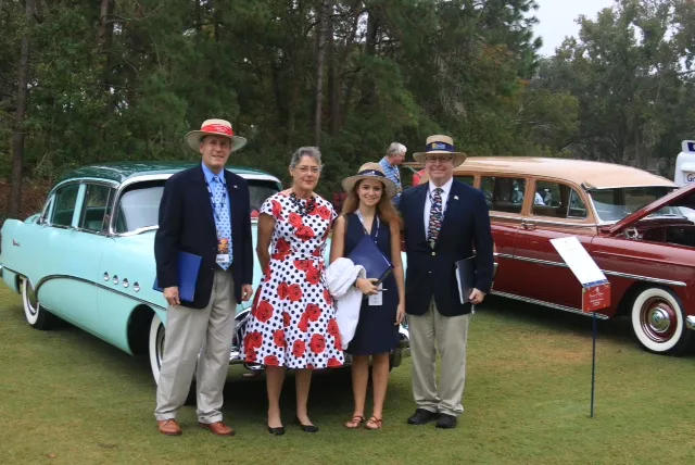 Four people in blazers, boater hats and dresses standing on a show-field lawn beside a seafoam-green sedan.