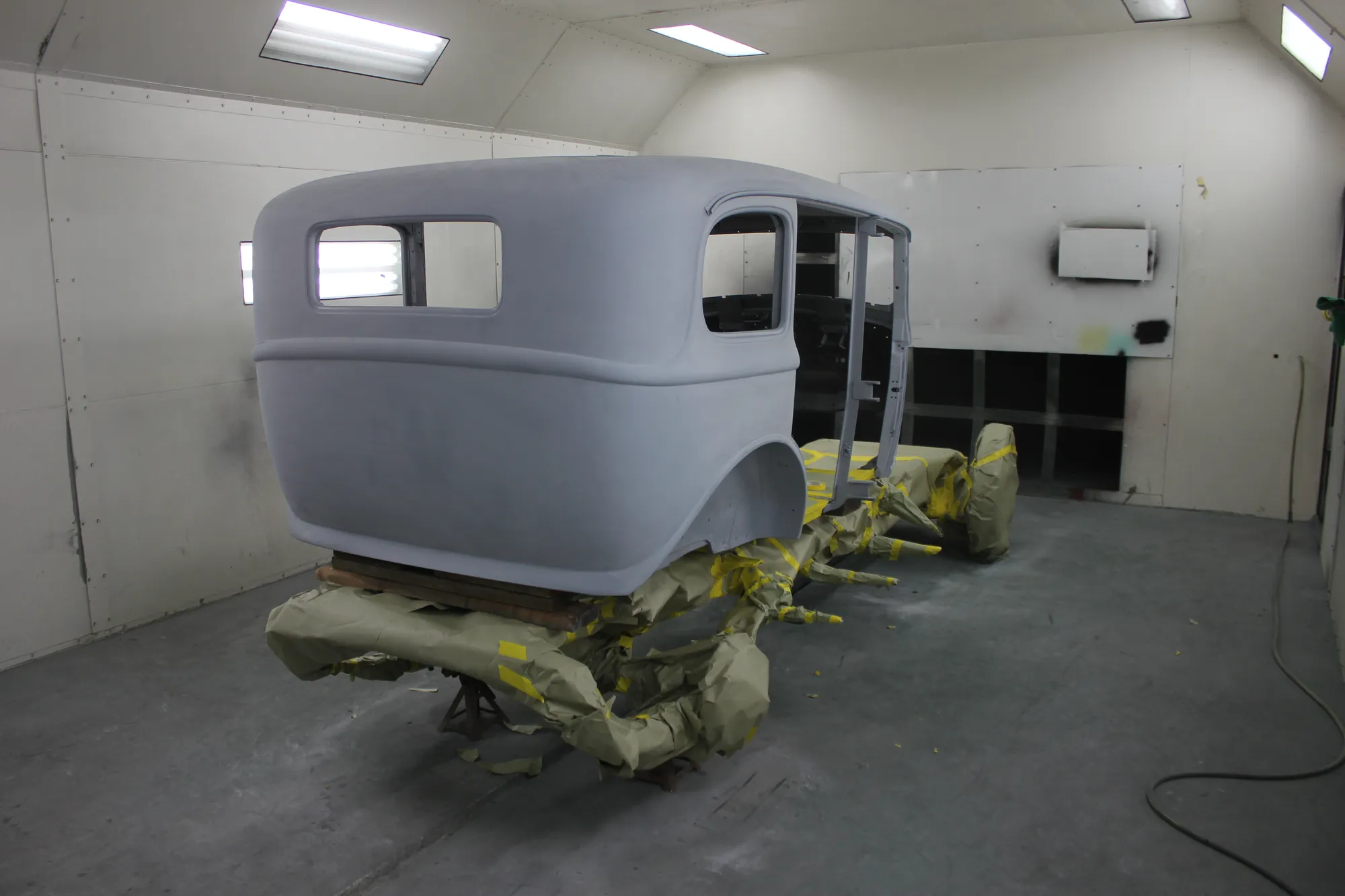 A primer-gray sedan body standing in the shop paint booth, masking paper and tape around its base.