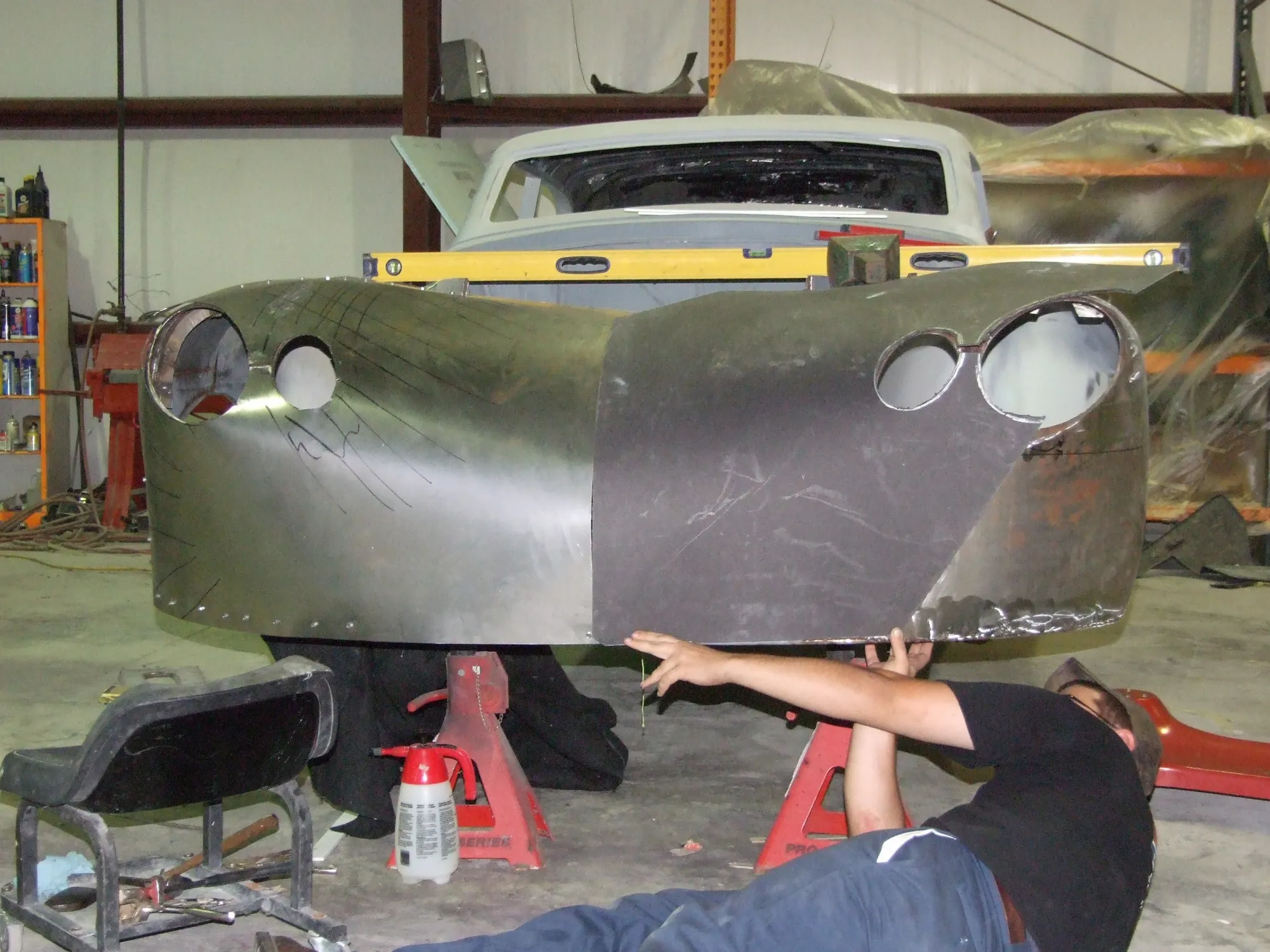 Hand-formed sheet metal fitted to a front end on stands, a worker on his back underneath it.