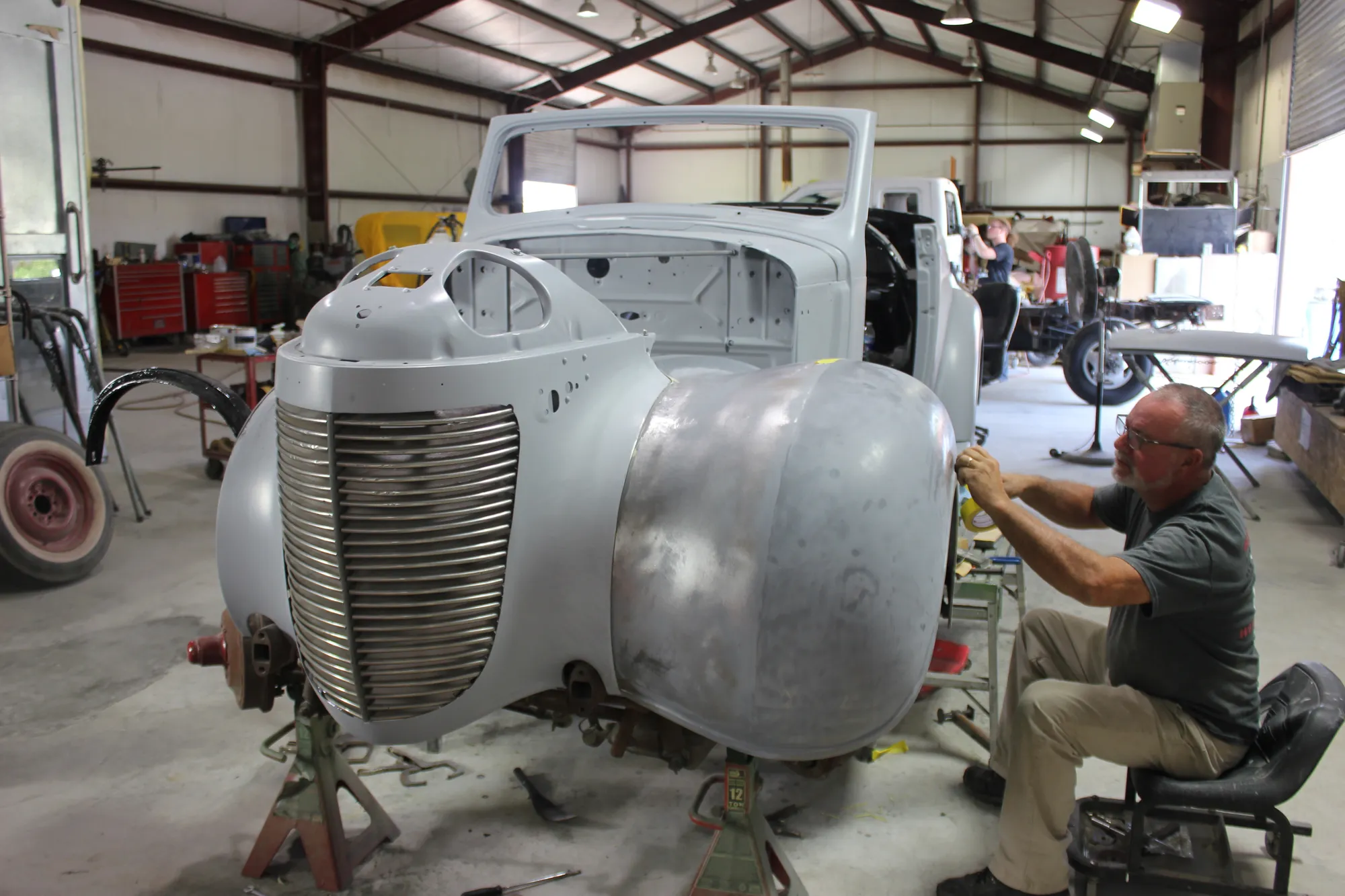 A GYR craftsman hand-sanding a bare-metal, primer-grey classic body on jack stands in the shop